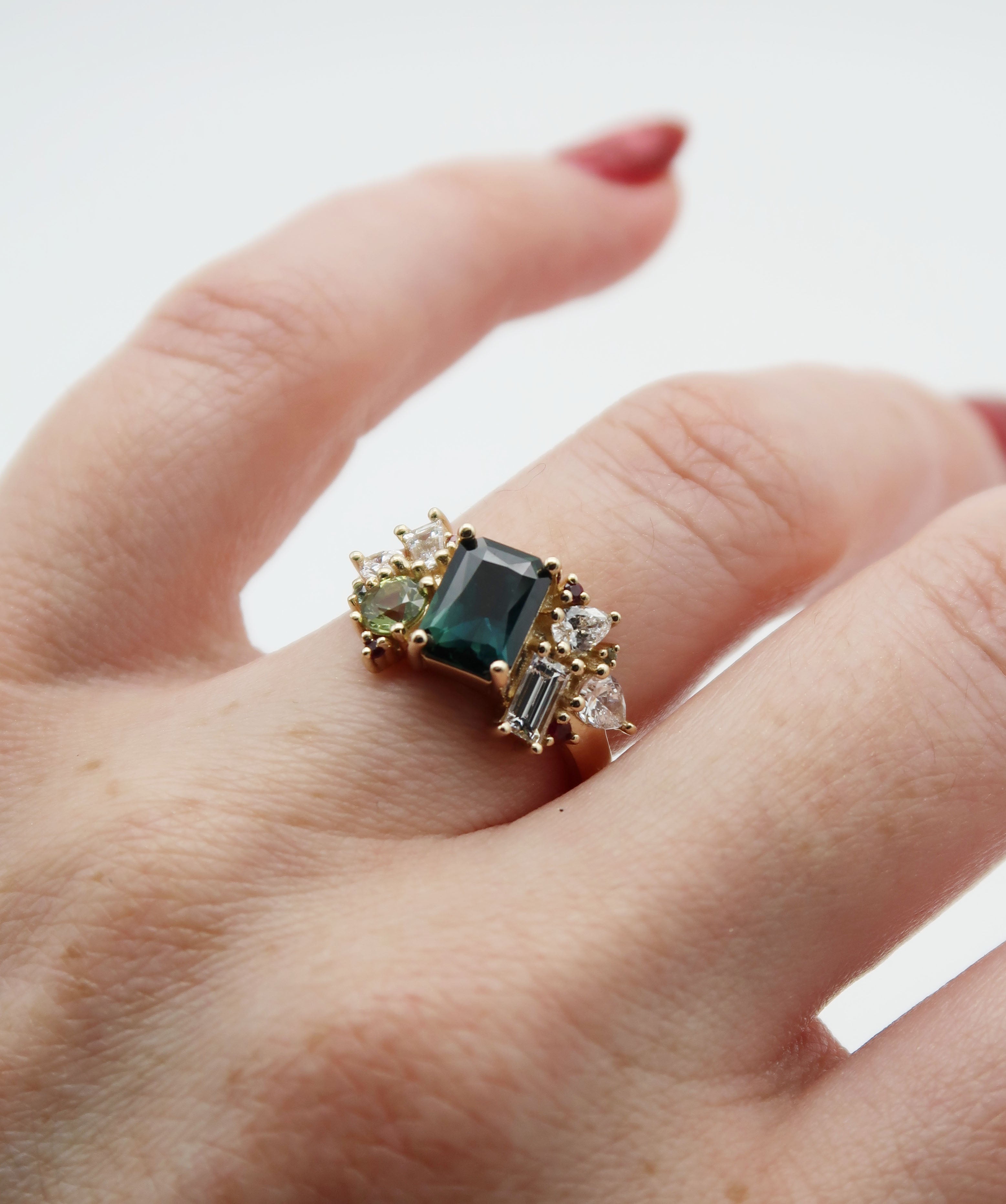 Bespoke 14k yellow gold and Australian sapphire cluster engagement ring. This statement piece features a mesmerizing deep blue centre stone, flanked by asymmetric clusters of green sapphires, diamonds and rubies, inspired by a bouquet of wild flowers. 