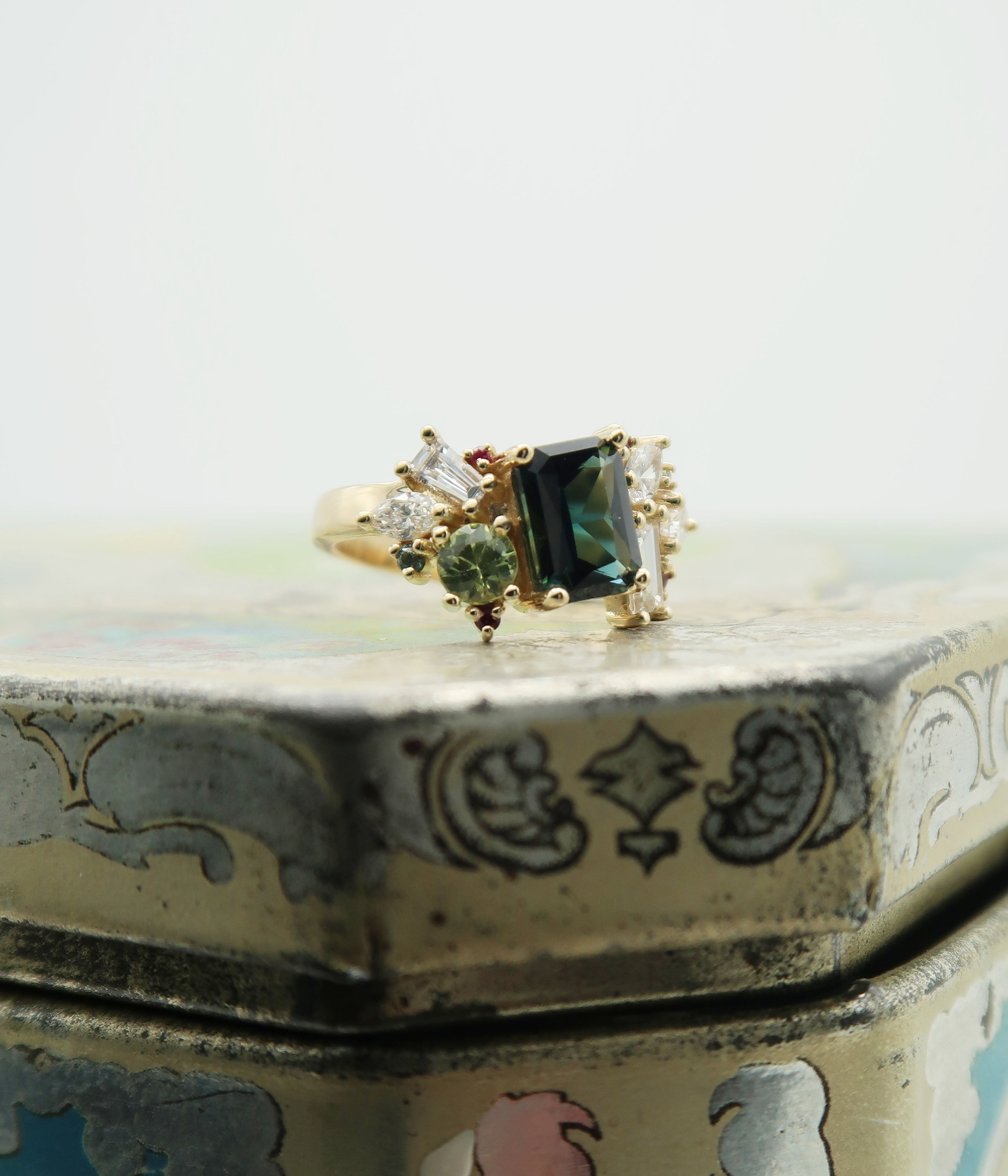 Bespoke 14k yellow gold and Australian sapphire cluster engagement ring. This statement piece features a mesmerizing deep blue centre stone, flanked by asymmetric clusters of green sapphires, diamonds and rubies, inspired by a bouquet of wild flowers. 

