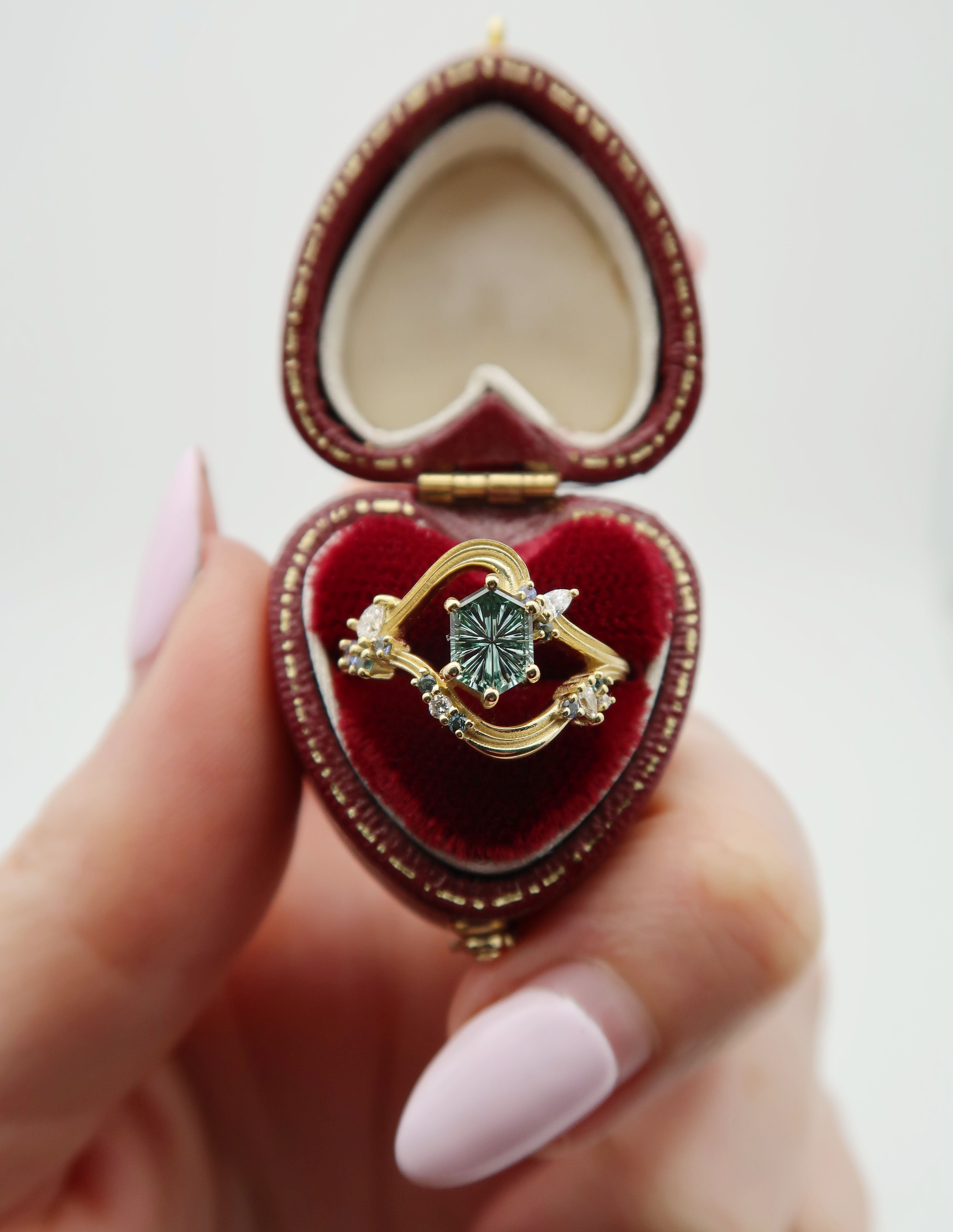 Custom engagement ring design with gold wedding band, blue gemstones, and diamonds on a heart-shaped red velvet ring box 