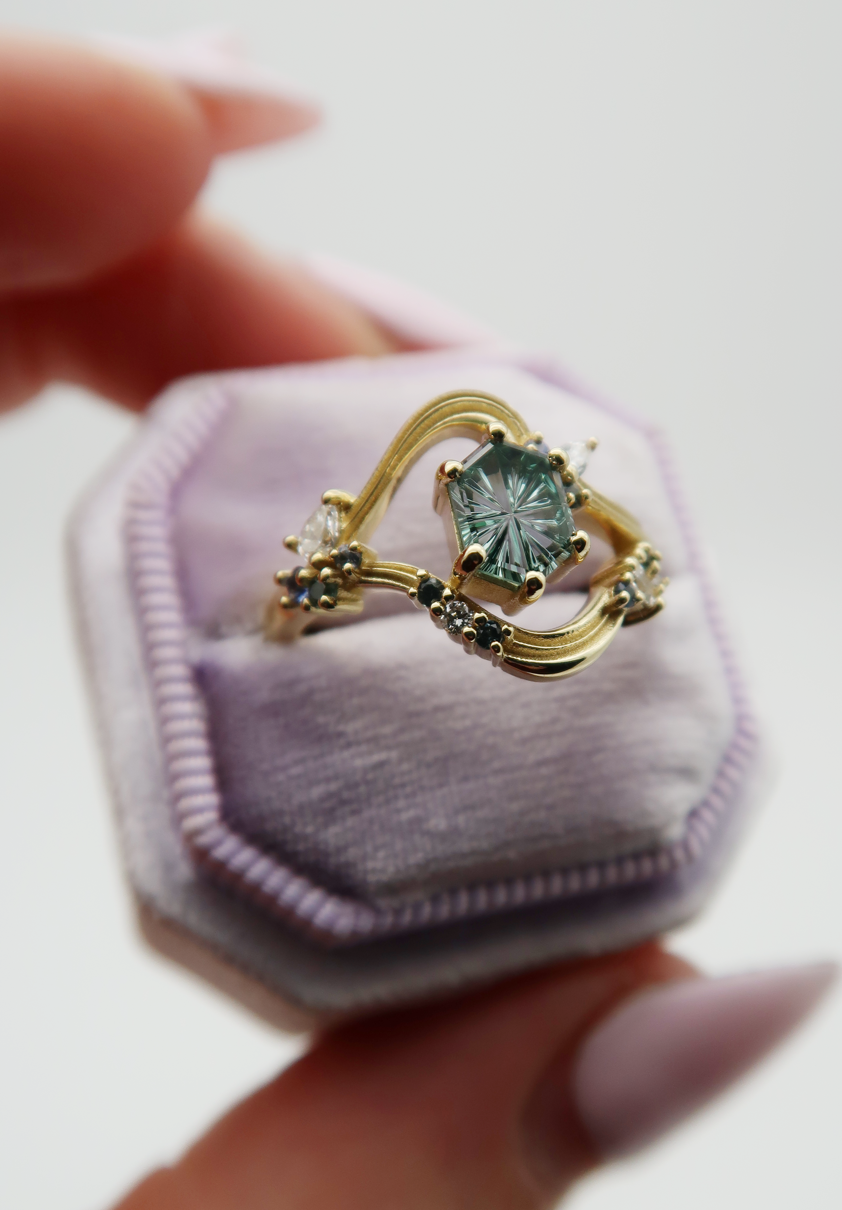 Custom golden engagement ring design with blue center stone surrounded by smaller blue gemstones and diamonds on a lavender velvet ring box.
