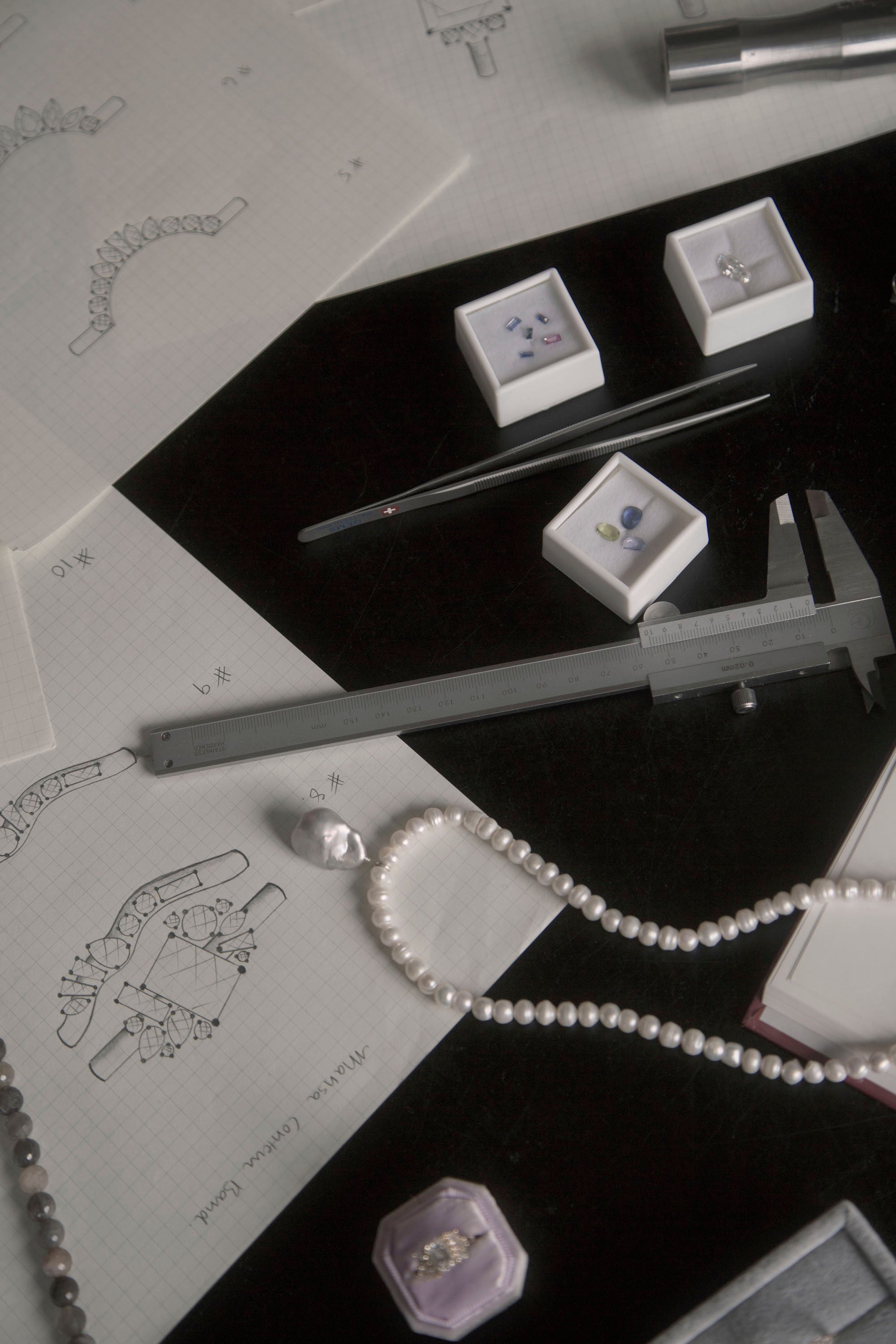 Black table with grid paper and custom ring design sketches, jewelry design tools, and loose gemstones on Invidiosa Jewellery's studio table.