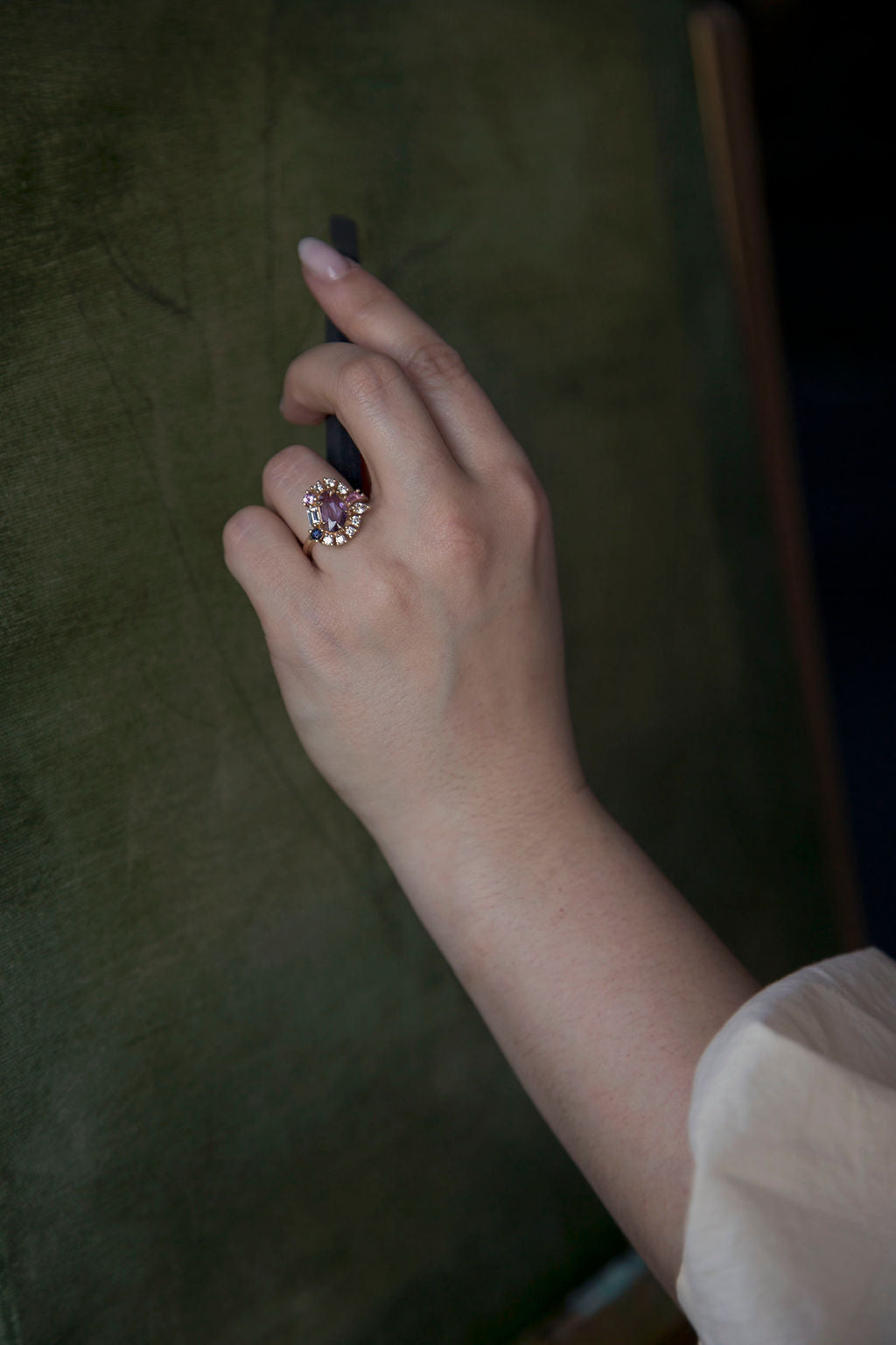 A woman's hand using a charcoal stick on a green painting canvas while wearing a custom engagement ring on a gold band with a purple center gemstone surrounded by a halo of small diamonds. 