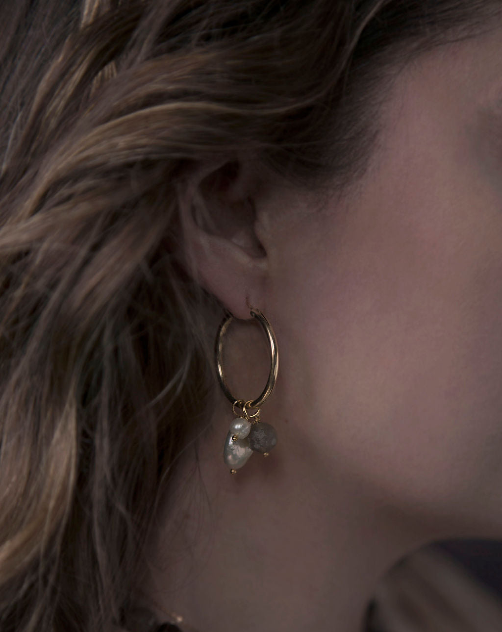 Close-up of a woman with brunette hair wearing a medium-sized gold hoop earring with three pearl charms dangling off the bottom. 