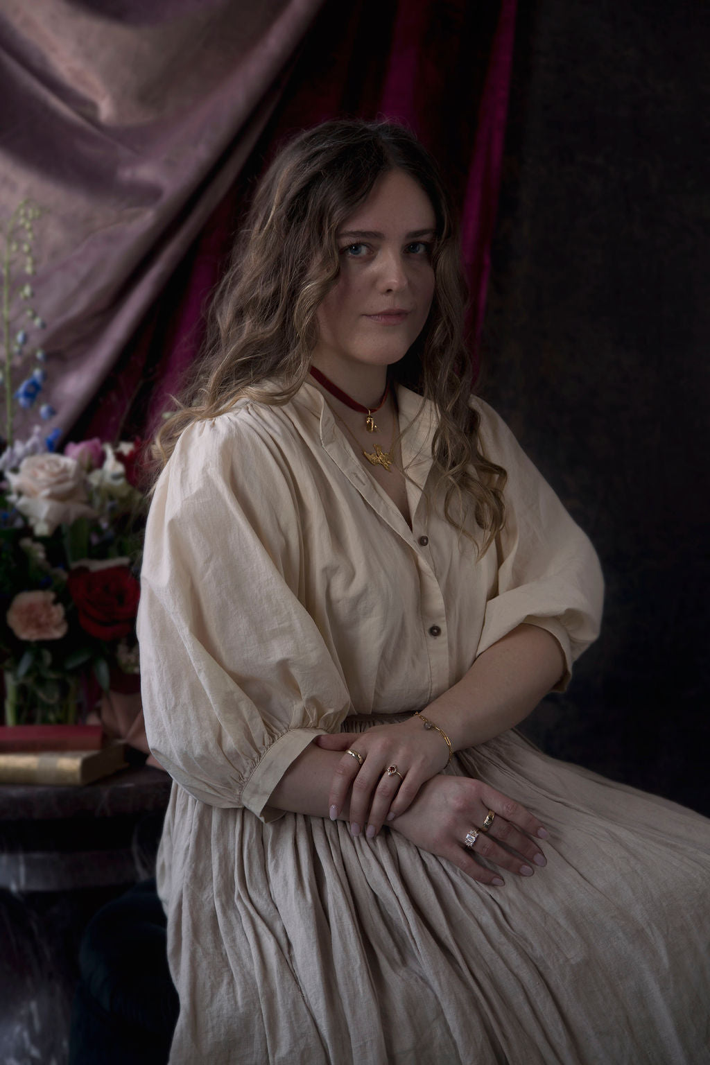 Brunette woman in a beige linen dress posing for a fine art portrait with her hands clasped in her lap, wearing four bespoke rings, a golden bracelet, and two bespoke necklaces by Invidiosa Jewellery. 
