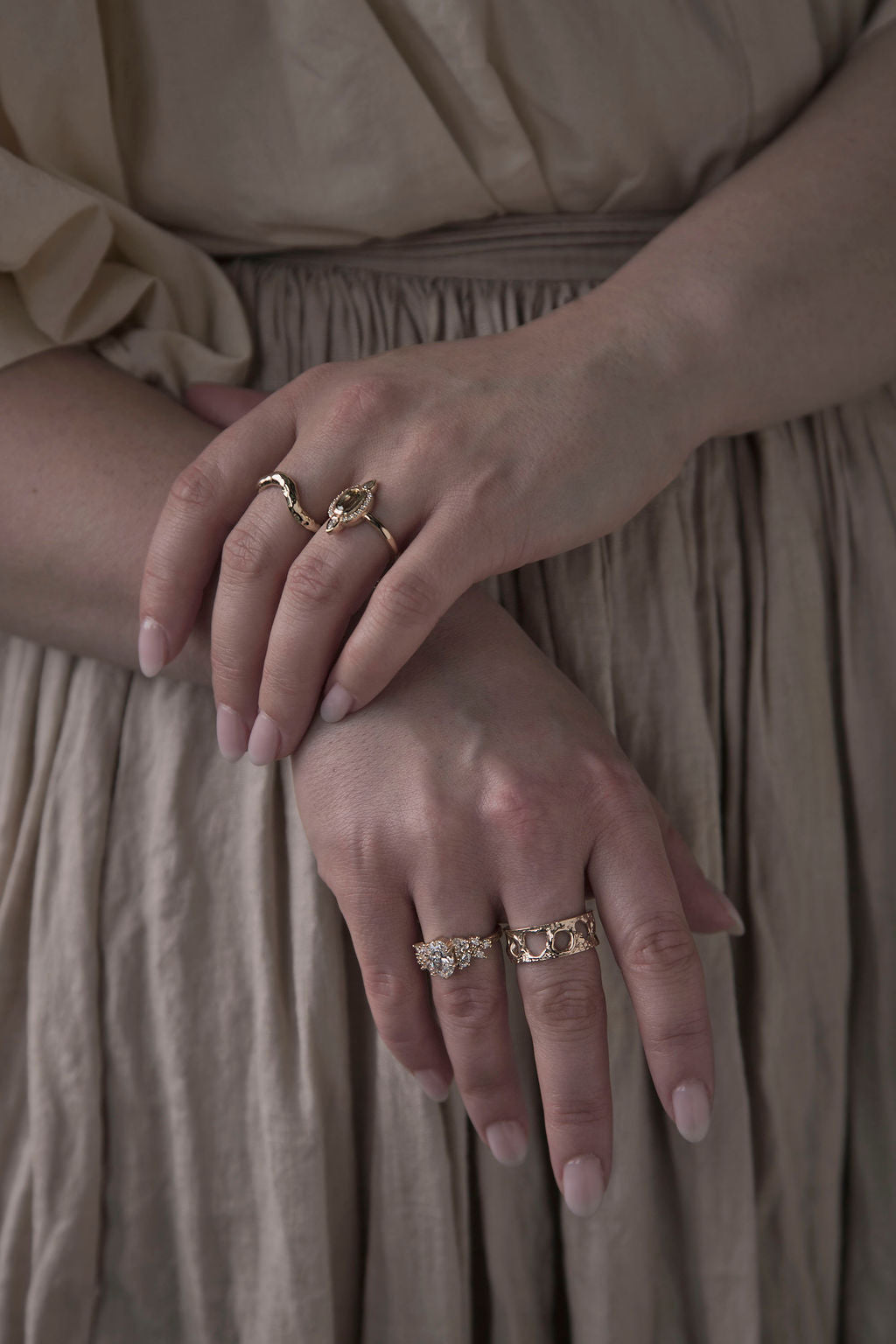 Woman in beige linen dress clasping her hands in front of her to show two bespoke gold wedding bands and two custom-designed engagement rings with large gemstones, accompanied by smaller diamonds. Each was handcrafted in their Toronto studio by Invidiosa Jewellery. 