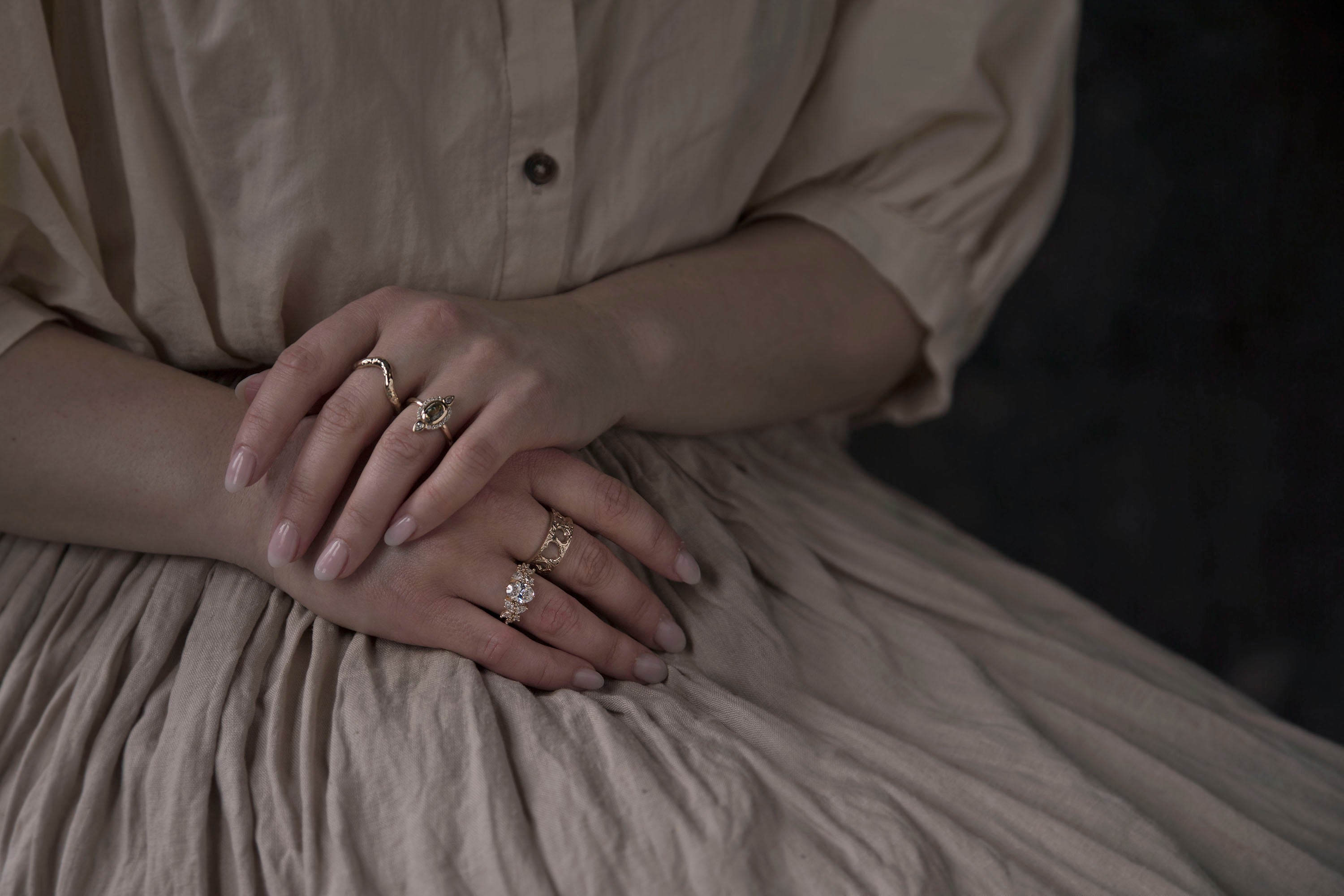Woman in beige linen dress with her hands clasped on her lap, while wearing two bespoke gold wedding bands and two bespoke gemstone engagement rings, all designed and handcrafted in Toronto by Invidiosa Jewellery. 
