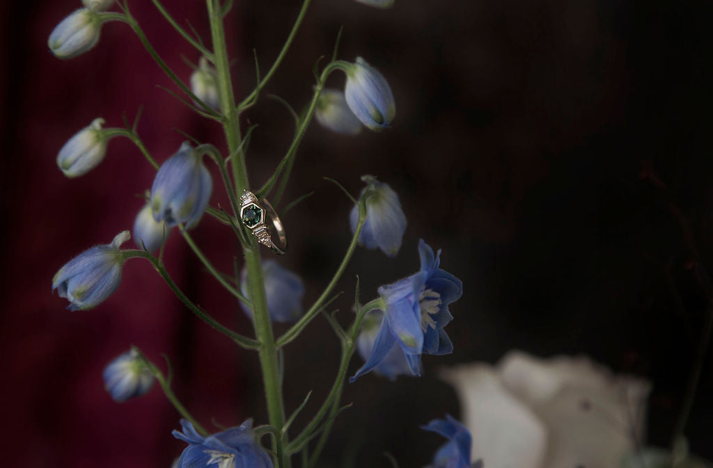 Bespoke custom engagement ring designed by Invidiosa Jewelry hanging off of a green and blue floral branch. 