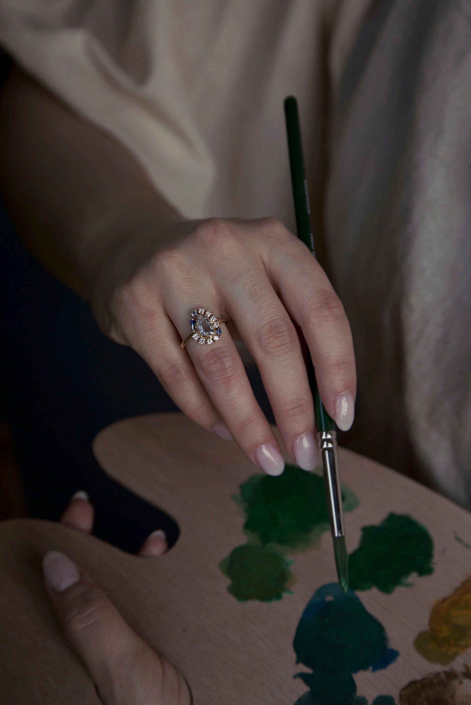 Woman painting while wearing a blue gemstone bespoke engagement ring surrounded by a three-quarter halo of small diamonds and gemstones set on a gold band. Ring was designed by Invidiosa Jewellery. 