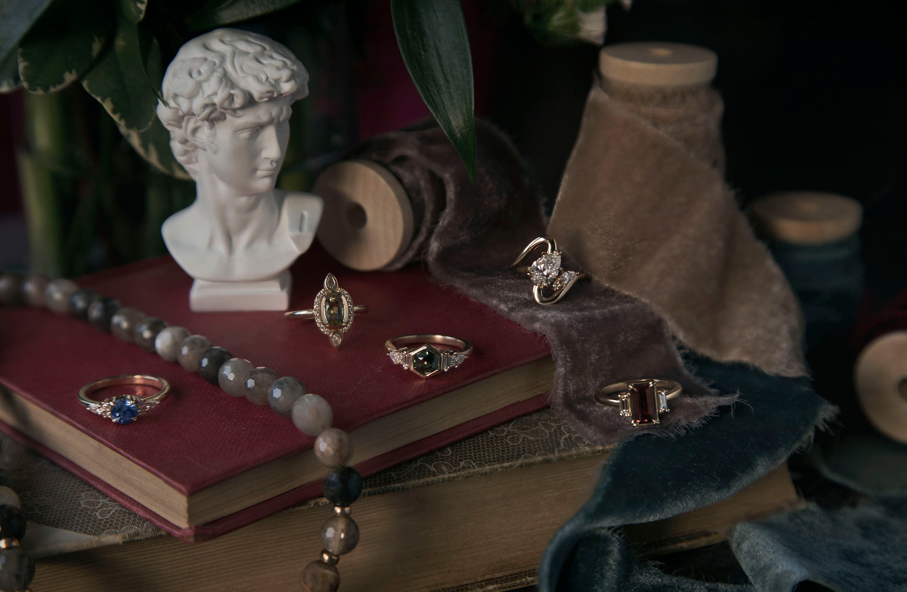 Vintage style still life with books and velvet ribbons showcasing five bespoke engagement rings with different colored gemstones and diamonds on gold bands. Each piece was designed by Invidiosa, a jewellery in their Toronto studio. 