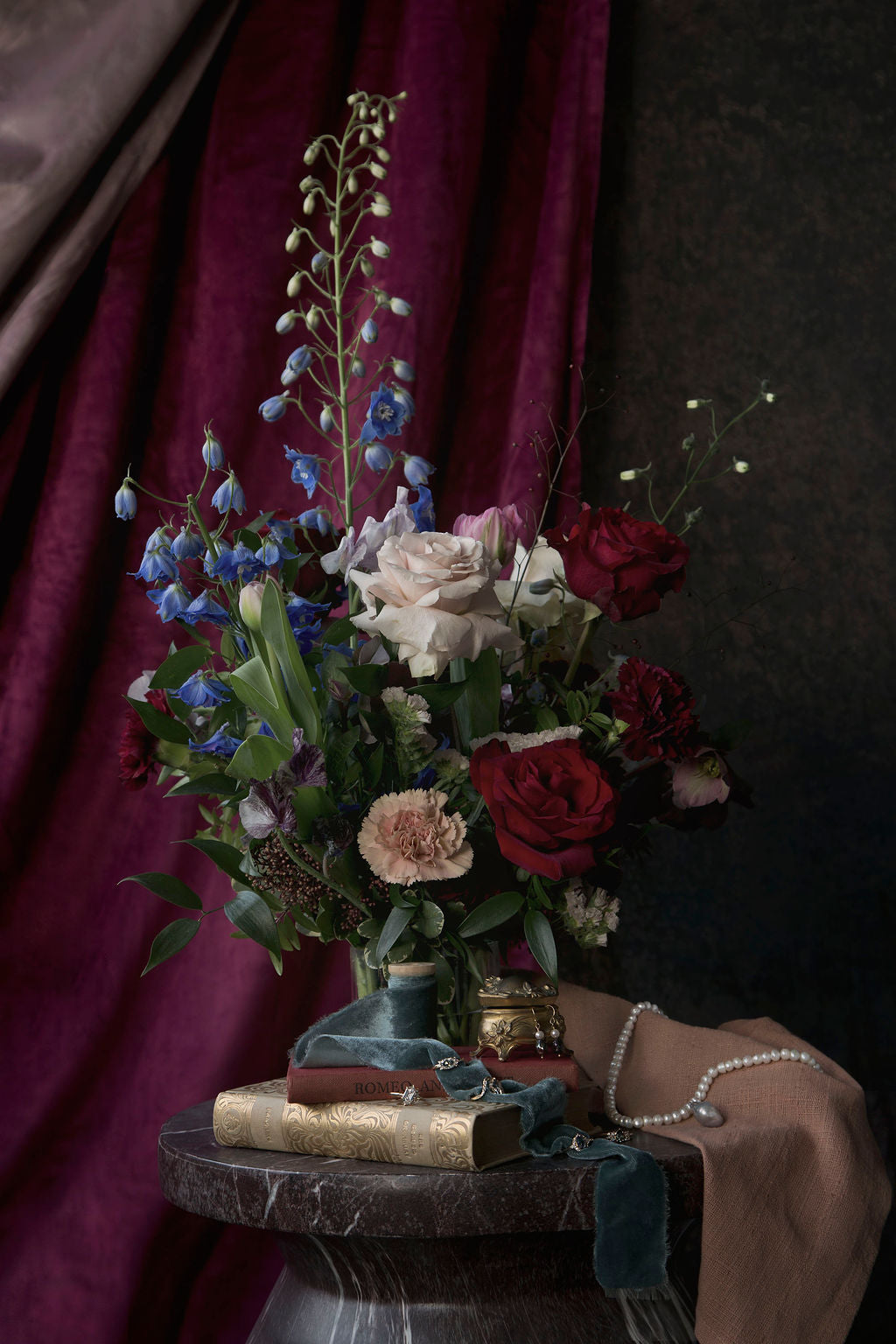 Poetic and painterly floral bouquet in a vase atop stacked books with gold trinkets and jewelry laying about, all sitting on top of a round marble table 