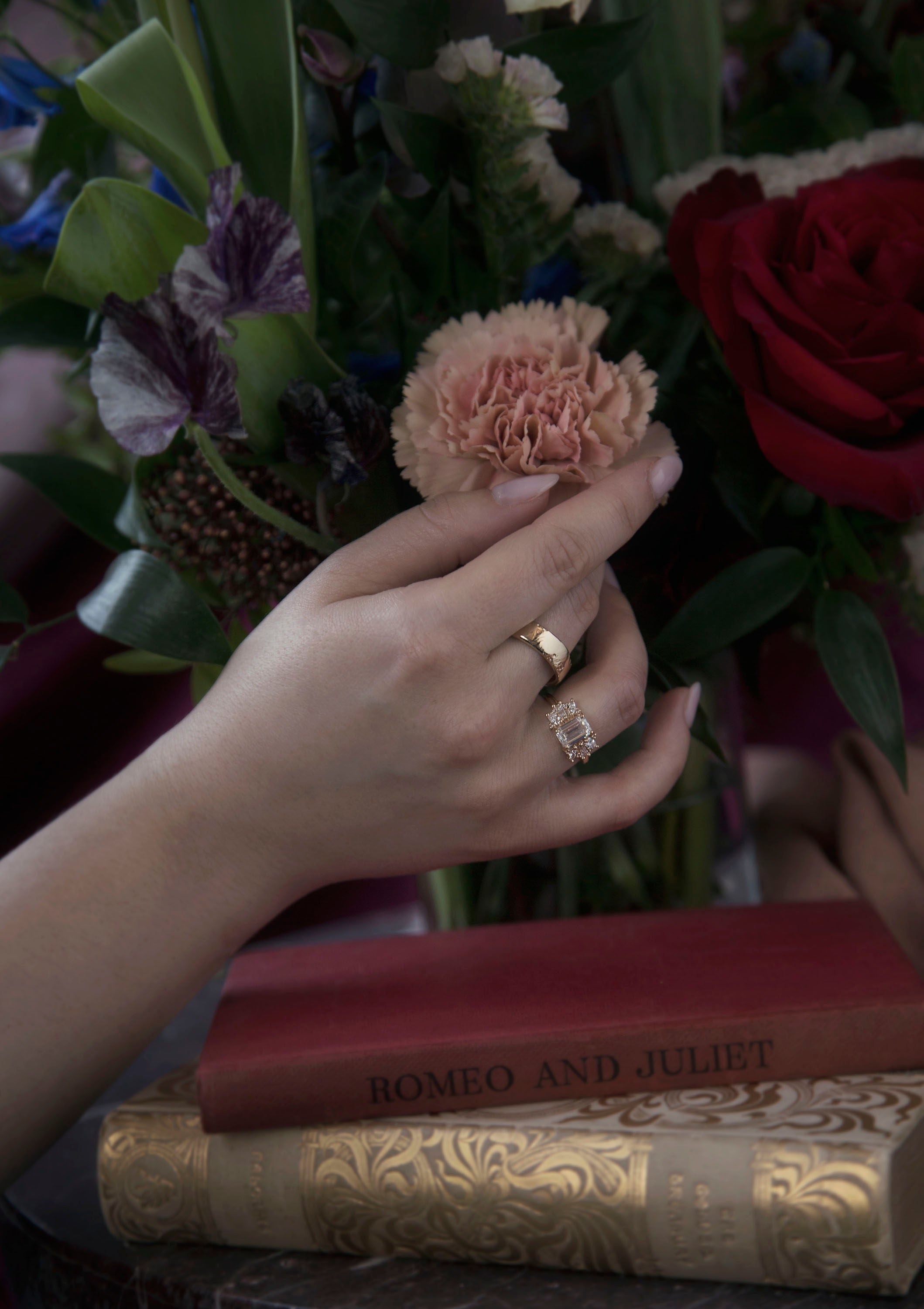 Woman's hand reaching for a floral bouquet while wearing a handcrafted golden wedding band accompanied by a bespoke diamond engagement ring. Each piece was designed by Invidiosa Jewellery. 