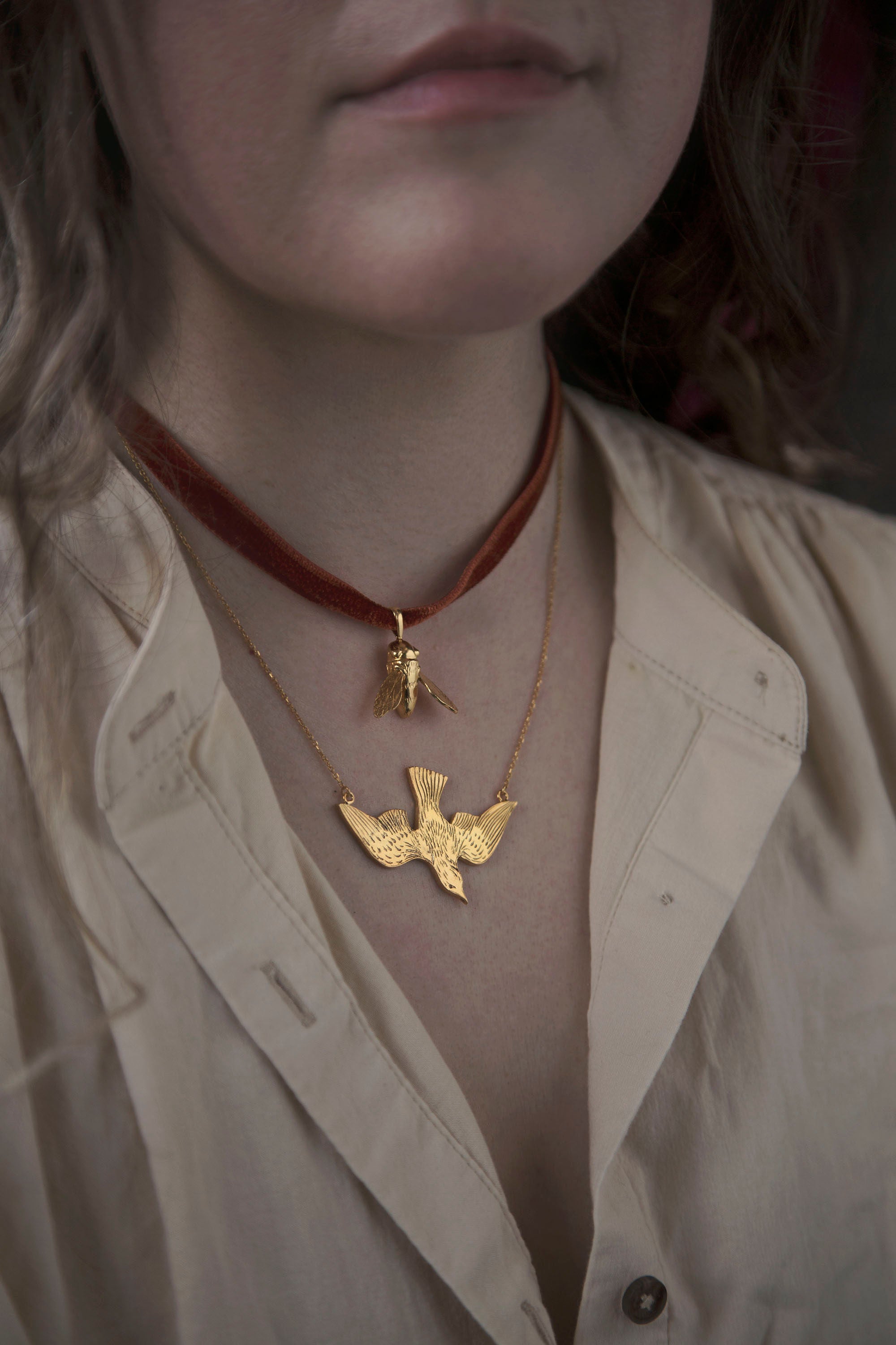 Brunette woman in a beige linen dress, wearing a velvet ribbon necklace with a golden bee pendant and a golden chain link necklace with a bird pendant. Each necklace was designed and hand-crafted in their Toronto studio by Invidiosa Jewellery. 
