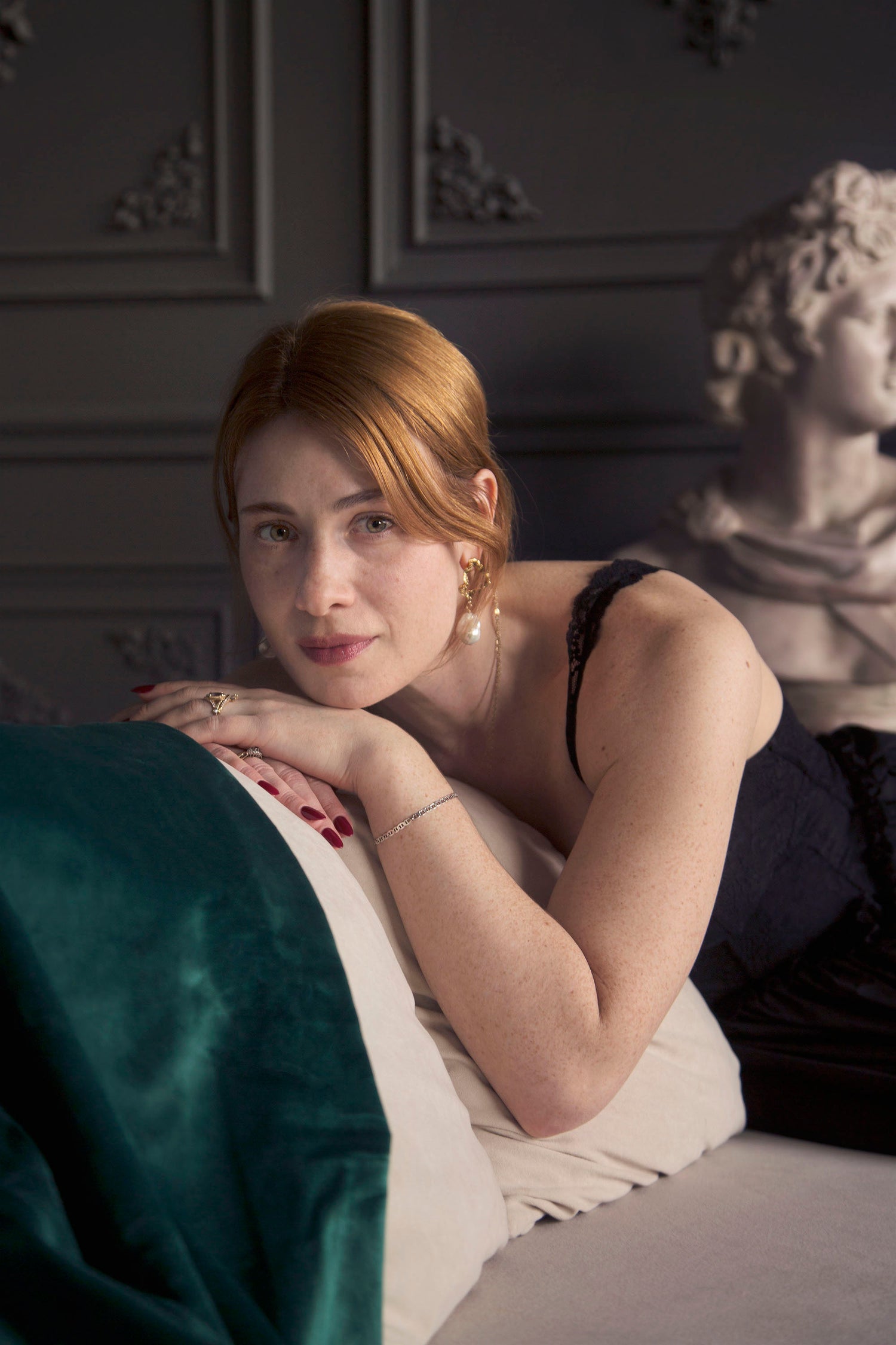 Portrait of founder of Invidiosa Jewellery, Tessa, in a black dress while lounging on a white couch in an estate-style room with a marble bust behind her. Tessa is wearing bespoke hand-crafted rings and earrings. 